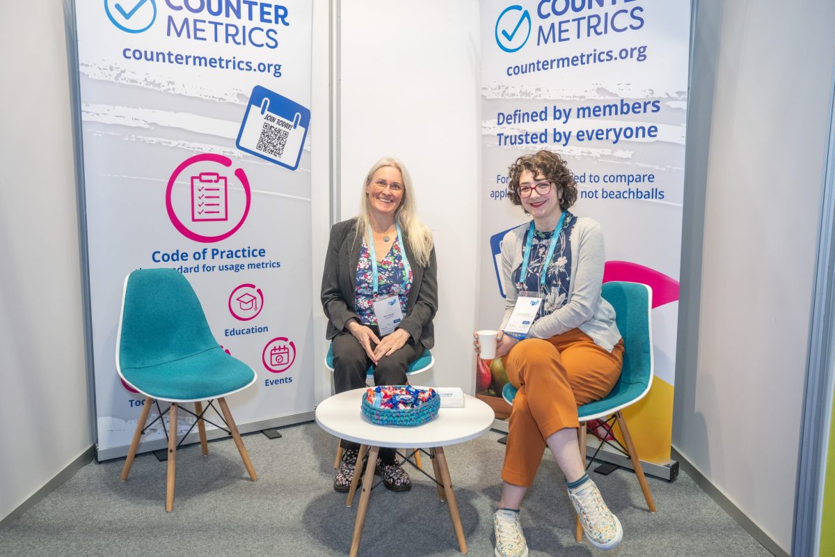 Siân (left), is a blonde woman wearing a grey suit and flowery top. She sits next to Tasha (right), wearing orange trousers and a flowery top. They are on the COUNTER stand at UKSG 2026, with two branded banners behind them and a small coffee table in front. Both are smiling.