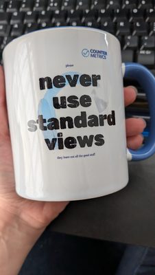 A white mug with blue handle. Printed on the mug is the phrase "never use standard views" in large black type, with "please" in tiny type above and "they leave out all the good stuff" in tiny type below.